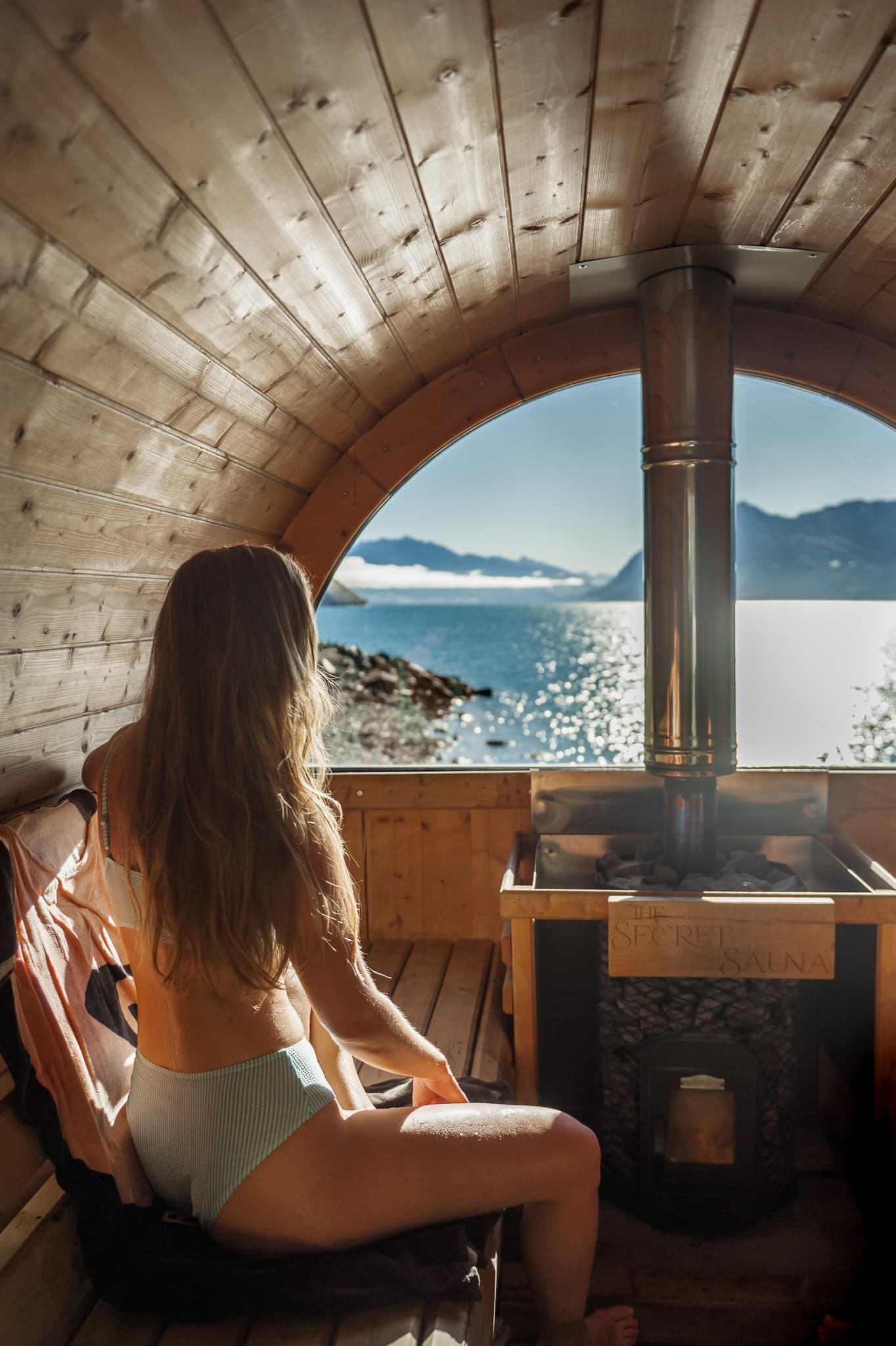 Woman enjoying lakeside views from inside a wooden sauna overlooking Lake Hāwea.